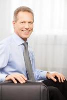 Ready to go. Smiling grey hair man in shirt and tie holding hand on suitcase while sitting on bed photo