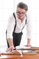 I need more information. Thoughtful young man in shirt and suspenders leaning at the table and reading a book laying on it photo