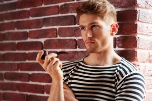 Sailor man. Handsome young man in striped shirt smoking pipe and looking away while leaning at the brick wall photo