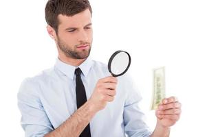 Fake or real Serious young man in shirt and tie looking through a magnifying glass at the paper currency while standing isolated on white background photo