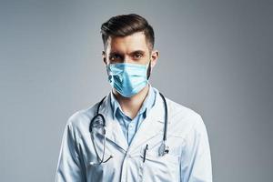 Confident young man in white lab coat and protective mask looking at camera while standing against grey background photo