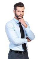 Thinking about solution. Thoughtful young handsome man in shirt and tie holding hand on chin and looking at camera while standing against white background photo