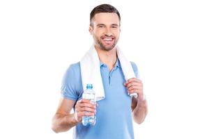 mantenerse hidratado. joven seguro de sí mismo con una toalla en los hombros sosteniendo una botella con agua y sonriendo mientras está de pie contra el fondo blanco foto