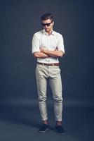 Casually handsome. Full length of confident young handsome man in sunglasses keeping arms crossed and looking down while standing against grey background photo