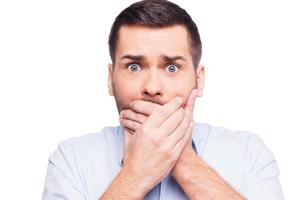 No way Shocked young man in formalwear covering mouth with hands and looking at camera while standing against white background photo