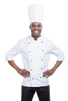 cocinar es mi vida confiado joven chef africano en uniforme blanco tomados de la mano en las caderas y sonriendo mientras está de pie contra el fondo blanco foto