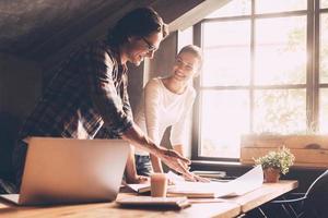 Working on project together. Cheerful young man and woman in casual wear pointing blueprint and communicating while both standing near wooden desk in creative office photo