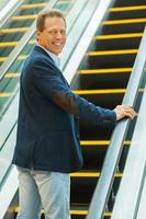 I am going up. Cheerful mature man looking over shoulder and smiling while moving up by escalator photo