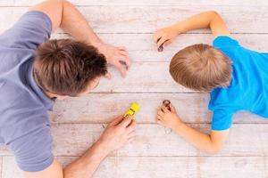 Playing together. Top view of father and son lying on the hardwood floor and playing with toy cars together photo