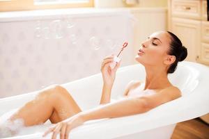 Bubble bath. Side view of beautiful young woman blowing soap bubbles while enjoying bubble bath photo