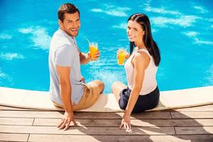 disfrutando de sus vacaciones de verano. vista superior de una pareja feliz con ropa informal sosteniendo vasos con jugo de naranja y sonriendo mientras se sientan juntos junto a la piscina foto