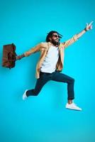 Full length of stylish young African man in casual wear smiling and carrying bag while hovering against blue background photo