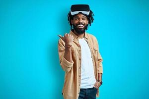 Happy young African man in virtual reality glasses pointing copy space and smiling while standing against blue background photo