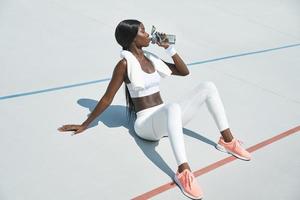 bella joven africana con ropa deportiva bebiendo agua mientras se sienta en una pista deportiva al aire libre foto