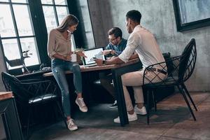 Three young people in smart casual wear analyzing data on laptop while working in the office photo