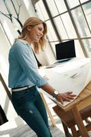 Busy young woman making a graph on the blueprint and smiling while working in the office photo