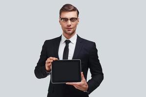 Copy space on his digital tablet. Good looking young man in full suit holding digital tablet and looking at camera while standing against grey background photo