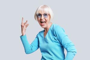 Rock it Playful senior woman making rock on sign and smiling while standing against grey background photo