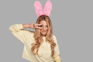 conejito gracioso. atractiva joven mujer sonriente con orejas de conejo rosa gesticulando y mirando a la cámara mientras se enfrenta a un fondo gris foto