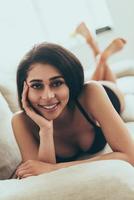 Flirty beauty. Beautiful young mixed race woman in black lingerie looking at camera and smiling while lying on couch at home photo