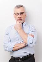 Thinking about your problem. Thoughtful senior man in formalwear holding hand on chin and looking at camera while standing against white background photo