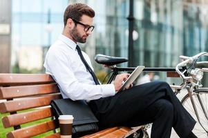 revisando su agenda de negocios. vista lateral de un joven hombre de negocios confiado que trabaja en una tableta digital mientras se sienta en el banco cerca de su bicicleta con un edificio de oficinas en el fondo foto