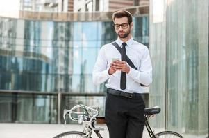 Sending quick message. Handsome young businessman holding mobile phone while leaning at his bicycle with office building in the background photo