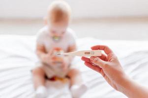Baby care. Hand holding thermometer while baby sitting on the bed photo