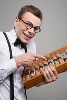 Old-fashioned calculator. Low angle view of cheerful young man in bow tie and suspenders holding abacus and pointing it while standing against grey background photo