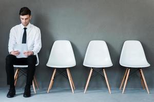 esperando entrevista. joven empresario confiado sosteniendo papel mientras se sienta en una silla con fondo gris foto