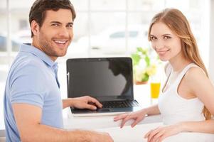 Surfing the net together. Handsome young man sitting at the table and using laptop while his girlfriend standing behind him photo
