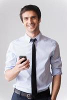 I am always in touch Confident young man in formalwear holding mobile phone and smiling while standing against grey background photo