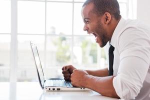 oficinista estresado. vista lateral de un furioso joven africano con camisa y corbata gritando mientras mira la laptop mientras se sienta en su lugar de trabajo foto