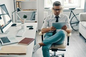 concentración total. un joven apuesto con camisa y corbata usando una tableta digital y sonriendo mientras está sentado en la oficina foto