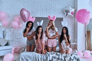 Oops...  Four playful young smiling women in bunny ears covering mouth with hand and looking away while sitting on the bed with balloons around photo