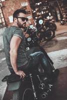 Freedom is in his veins. Rear view of confident young man sitting on his bike and looking over shoulder with motorcycle garage in the background photo