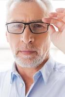 Confident look. Confident grey hair senior man in shirt adjusting his eyeglasses and looking at camera while standing in front of the window photo