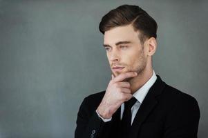 elegancia y confianza. un joven hombre de negocios pensativo sosteniendo la mano en la barbilla y mirando hacia otro lado mientras se enfrenta a un fondo gris foto