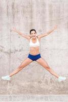 feliz de vivir en forma y saludable. hermosa joven sonriente estirando los brazos y las piernas mientras salta frente a la pared de hormigón foto