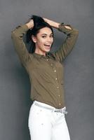 Feeling so excited. Happy young woman in smart casual wear standing against grey background and holding hands in hair photo