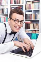 Computer nerd. Side view of excited young man in shirt and bow tie typing something on computer and looking at camera with smile photo