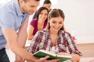 Students studying together. Two cheerful students discussing something while looking at the book together with other students sitting behind them photo