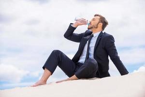 Refreshing his mind. Young businessman drinking water while sitting on the top of sand dune photo