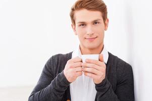 Relaxing with cup of fresh coffee. Handsome young man holding cup of coffee and looking at camera while leaning at the wall photo