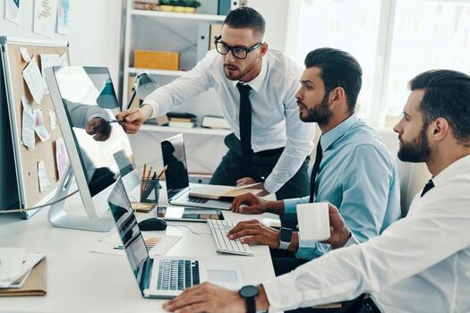 Developing new approaches. Group of young modern men in formalwear working using computers while sitting in the office photo