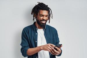 Happy young African man in casual wear listening music and smiling while standing against grey background photo