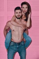 Feeling loved. Handsome shirtless man carrying his girlfriend on shoulders while standing against pink background photo