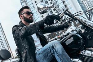 Heartbreaker. Handsome young man in leather jacket and sunglasses riding motorbike while spending time outdoors photo