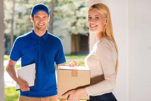 First class delivery service. Beautiful young woman holding a cardboard box while young delivery manholding clipboard and smiling photo
