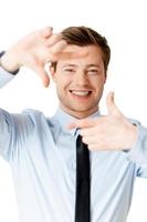 centrándose en ti. apuesto joven con camisa y corbata enfocándose en ti con marco de dedo y sonriendo mientras está de pie aislado en blanco foto